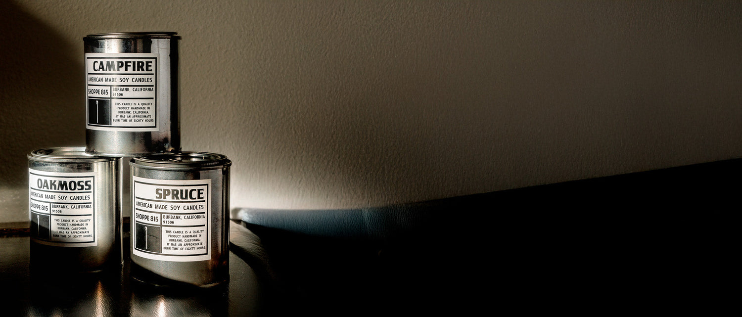 Shoppe 815_Tin Candle Collection - A stack of best selling candles, campfire, oakmoss, and spruce, lit by the sun in a dark room