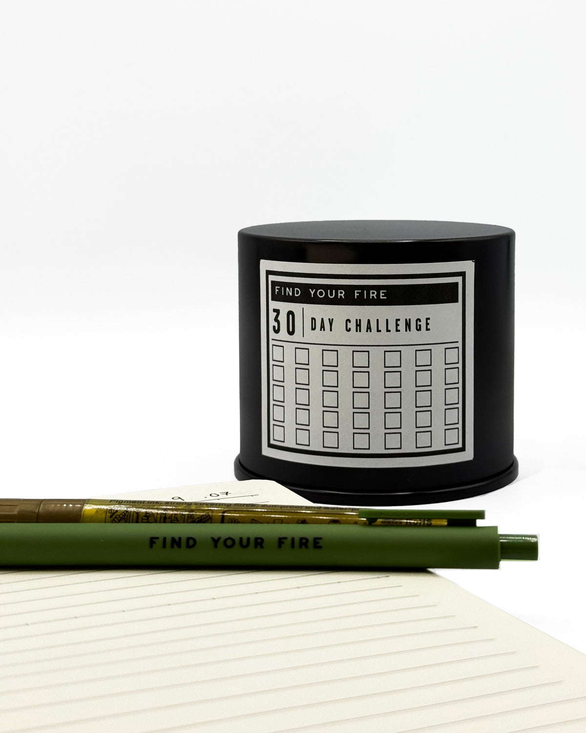 A black cylindrical container labeled 'FIND YOUR FIRE 30 DAY CHALLENGE' sits behind a journal dated 9.07 with a pen and gold marker resting on lined paper.