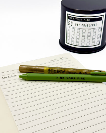 A green pen that says 'FIND YOUR FIRE' and a gold marker, resting on a lined notepad labeled 'DAY 1'. A black cylindrical candle container in the background features a '30 DAY CHALLENGE' grid and the same text.